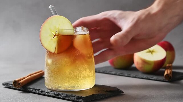 Hand places chilled apple cider cold brew tonic on slate board beside apples and cinnamon sticks.