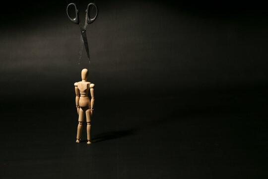 Wooden mannequin standing beneath a large pair of scissors pointing down at its head, symbolizing looming danger, hidden threats, or impending cuts.