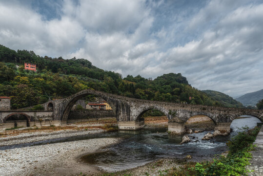 View of the historic Devil's Bridge spanning the Serchio river, surrounded by lush green hills and a cloudy sky in Borgo a Mozzano, Tuscany, Italy.