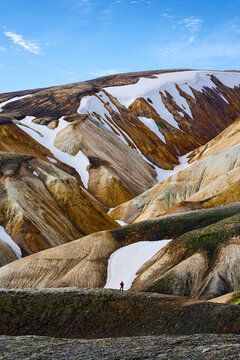 View of Landmannalaugar with a lone hiker in a red jacket standing amidst colorful rhyolite mountains and patches of snow in Rangarthing ytra, Iceland.