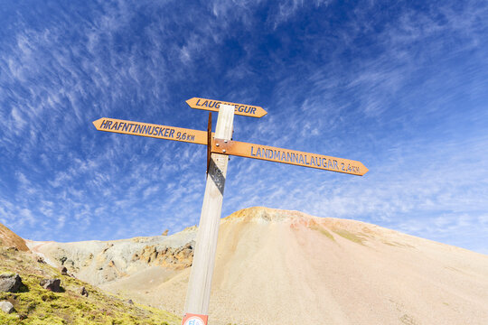 View of a wooden hiking trail signpost in Landmannalaugar with directions to Laugavegur and Hrafntinnusker against colorful volcanic mountains under a blue sky in Rangarthing ytra, Iceland.