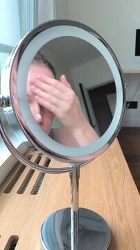 Applying green concealer base on face skin looking into a makeup mirror. Young woman masks redness and color corrects sensitive epidermis areas at home. Mirror reflection captures