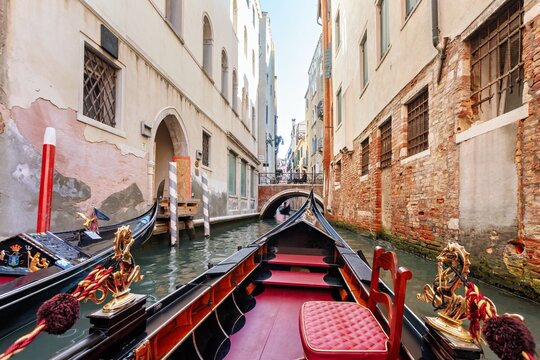 View of a traditional gondola navigating a narrow canal in Venice, with historic buildings, a small stone bridge in the background, and clear daylight, Venice, Veneto, Italy.