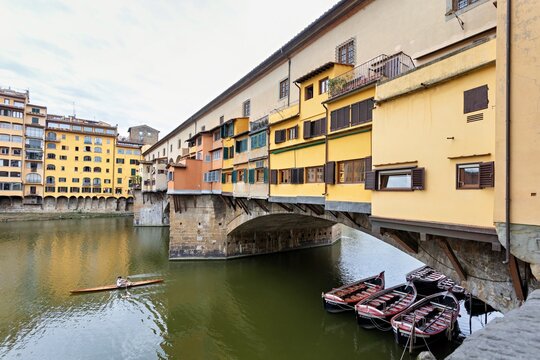 View of the historic Ponte Vecchio bridge with its iconic yellow buildings over the Arno River, featuring moored wooden boats and a person rowing in Florence, Tuscany, Italy.