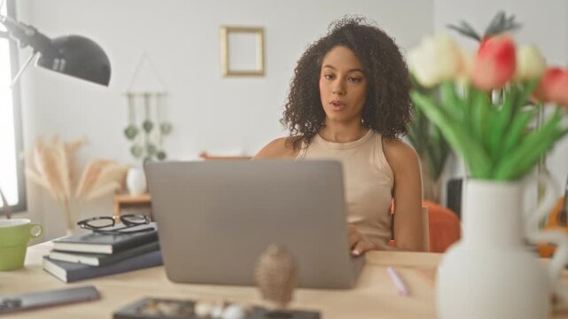 Woman with curly hair at laptop tilting head back and stretching neck in studio workspace, reaching for device; tired rest.