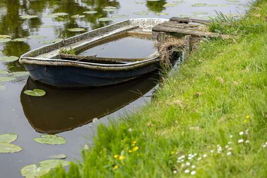 View of a small, partially submerged blue boat tied to a weathered wooden dock on a canal bank with green grass and lily pads in Monnickendam, North Holland, Netherlands.