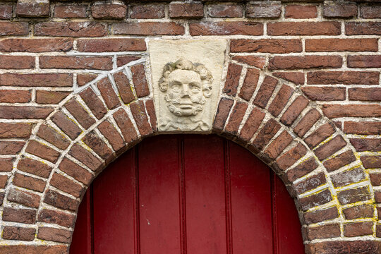 View of a carved stone face keystone above a red wooden door set in a brick archway, Monnickendam, North Holland, Netherlands.