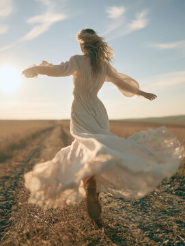 Carefree young woman in flowing white bohemian dress twirling in open field at sunset, backlit full body view, freedom and summer countryside outdoor lifestyle stock photo, joyful girl spinning field