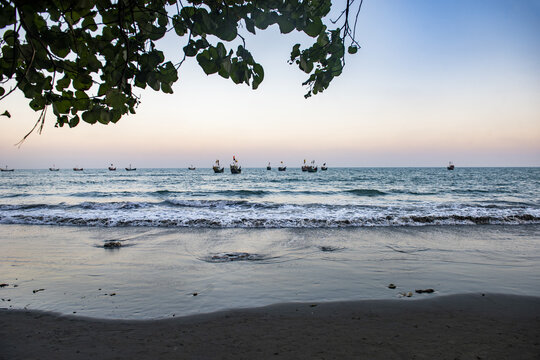 View of Saint Martins Island with fishing boats on the sea under a twilight sky, framed by tree branches on the sandy beach in Cox's Bazar District, Chittagong Division, Bangladesh.