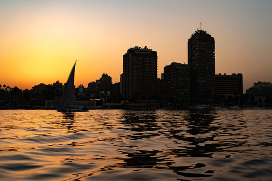 View of a traditional felucca boat sailing on the Nile River during a golden sunset with city skyline silhouettes in Maadi as Sarayat Al Gharbeyah, Cairo Governorate, Egypt.