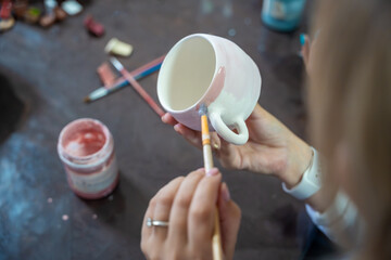 Woman painting pink glaze on handmade ceramic mug with brush in pottery workshop. Pottery decoration represents creativity and learning a craft as a hobby © dtatiana
