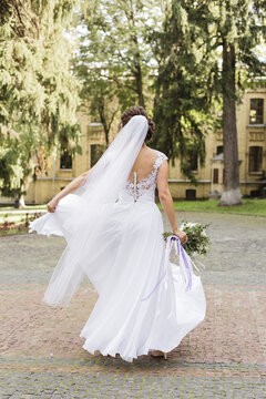 Happy bride in fluttering white dress and long veil twirling joyfully outdoors on cobblestone path.
