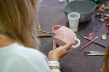Woman painting pink glaze on handmade ceramic mug with brush in pottery workshop. Pottery decoration represents creativity and learning a craft as a hobby © dtatiana