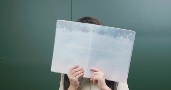 Playful Asian schoolgirl hiding face behind folder and smiling at camera