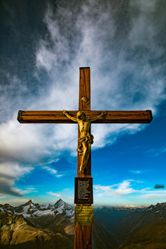 View of a wooden crucifix with a figure of Christ set against a dramatic sky with clouds over the snow-capped peaks of the Swiss Alps in Zermatt, Valais, Switzerland.