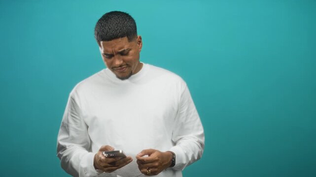 Man holding smartphone, frowning and rubbing forehead while reading messages on phone against a teal studio backdrop; frustration technical issue.