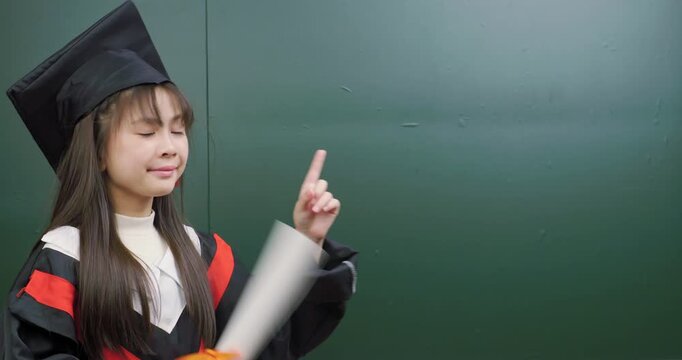 Cute child graduate pointing index finger up and to the side against background.