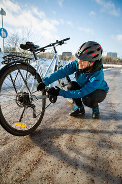 A mid-adult woman checks her bicycle chain on an icy road in Helsinki, Finland, demonstrating winter cycling maintenance and bike repair.