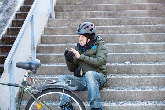 A senior man uses his smartphone while sitting on stairs beside his bicycle in Helsinki, Finland, resting during his winter commute.