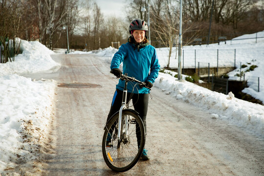 A smiling mid-adult woman stands with her bicycle on an icy path in a snowy Helsinki park, Finland, during winter commuting