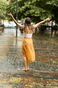 Back view of a carefree woman dancing barefoot on a wet paved sidewalk during heavy summer rain with hands wide open, feeling joy, freedom, and emotional release outdoors.
