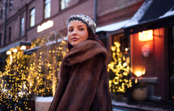 Young woman with dark hair wearing a fur coat and sequined headband stands outdoors in winter, illuminated by warm lights from nearby buildings and trees