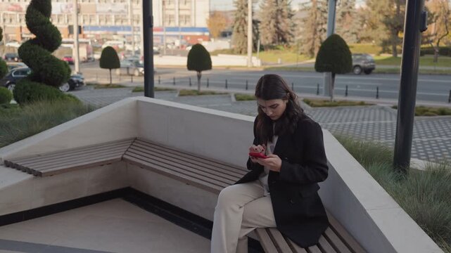 Focused young woman using smartphone while sitting on urban bench