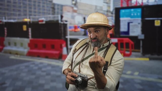 Man middle aged with grey beard and pith helmet shows middle finger while holding a vintage camera and backpack on a street construction site; defiance.