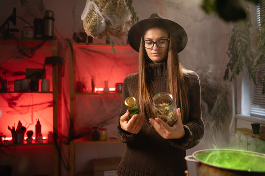 A realistic witch brewing a potion in a cauldron. holds a jar of ingredients and is selecting the one she needs. Everyday life intertwined with traditional witchcraft, magic, and a mystical atmosphere
