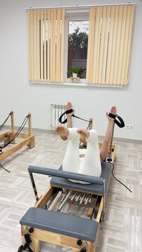 Reformer pilates workout showing legs lifted high while using black straps in a bright modern gym for core physical wellness training