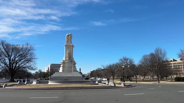 Wide tracking view of the Peace Monument beside a broad Washington DC avenue. Leafless trees and surrounding buildings frame the marble monument beneath a bright blue sky.