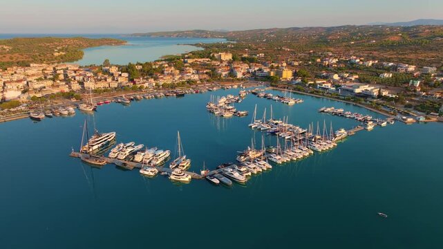 Aerial view of Porto Heli's calm harbor in Greece, with sailboats scattered across the water, waterfront hotels, and the peaceful town glowing in golden light.