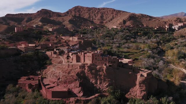 Elevated aerial shot showcasing an authentic earthen fortress and defensive citadel perched atop a dramatic rock cliff overlooking the Todra river valley in Morocco.