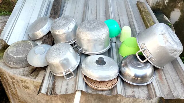 Various aluminum cooking pots and colorful plastic basins drying outdoors on a corrugated metal surface in a rustic village setting