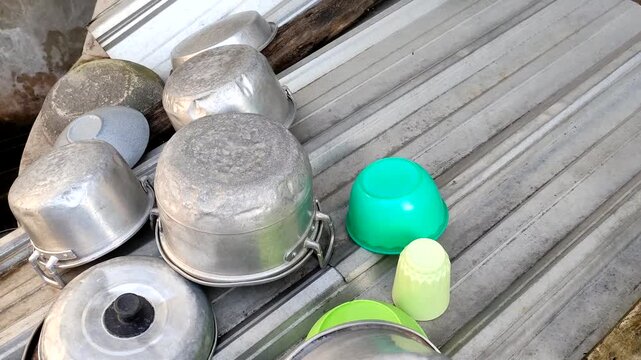 Close up of clean cooking pots, plastic bowls, and drinking cups upside down drying on a metal corrugated surface outdoor, traditional household kitchenware organized after washing.