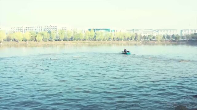 Fisherman in small boat casting net on calm river with urban buildings and trees in background