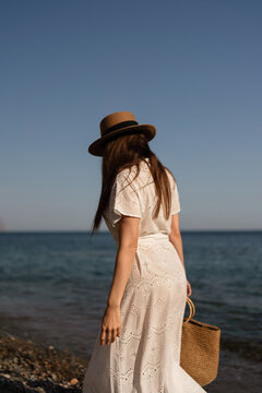 Woman beach vacation, long-haired female in hat and white dress walking along tranquil seashore with copy space
