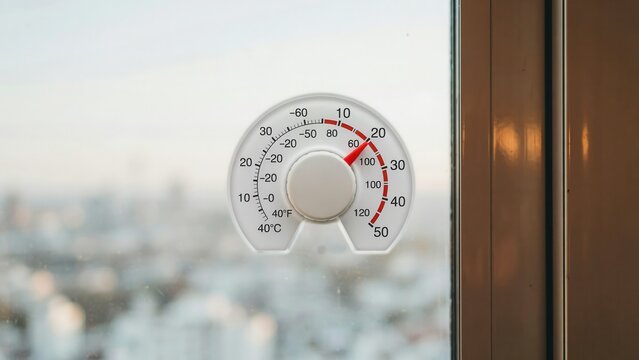 A circular window thermometer attached to a glass pane showing outdoor temperature against a blurred city background. Useful for weather and climate change editorial illustrations.