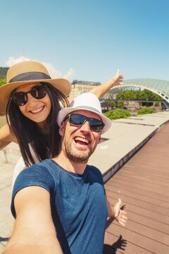 Happy young couple taking selfie in front of rike bridge in Georgia capital Tbilisi in summer. Two tourists having fun on romantic vacation - Holidays and traveling lifestyle concept.Weekend get away