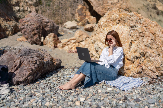 Young woman working on laptop while sitting on sunny pebble beach near large coastal rocks