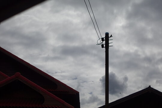 composition featuring a wooden utility pole with power lines, flanked by traditional red-tiled roof gables, set against a heavy, overcast cloudy sky. The scene captures a quiet residential neighborhoo