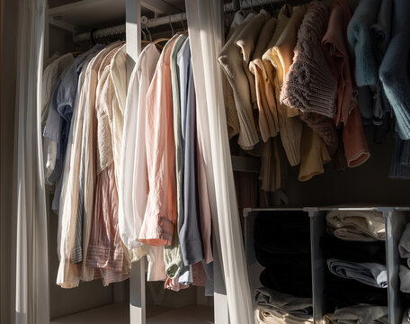 Sunlit open wardrobe filled with aesthetic pastel clothes and cozy knit sweaters, organized home interior with warm natural morning light.