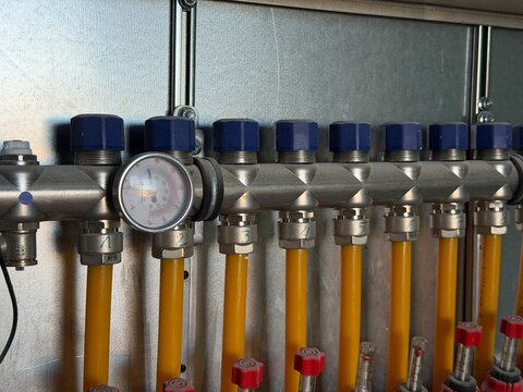 Heating manifold with brass valves and blue caps, pressure gauge and yellow flexible pipes controlling warm water flow and temperature for underfloor radiant heating systems