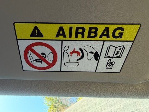 Airbag warning label on a car sun visor showing pictograms and text advising no rear facing child seats in front, proper occupant positioning and consult owner's manual