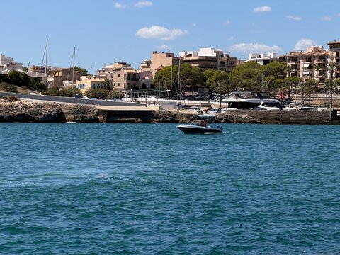 Motorboat navigating tranquil blue waters along the rocky coastline of a bustling mediterranean town in manacor, balearic islands, with buildings and a marina under a clear sky
