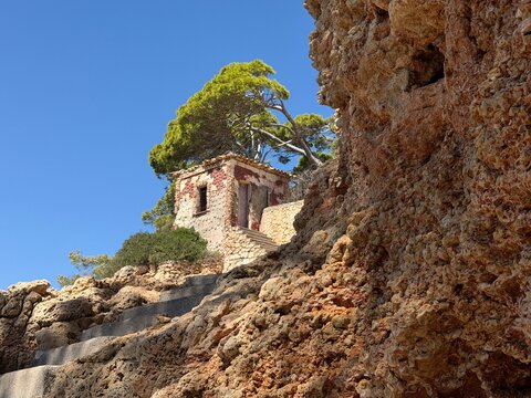 Old stone house built into a rugged rocky cliff near manacor, balearic islands, with weathered facade, winding concrete steps, sparse mediterranean vegetation and clear blue sky