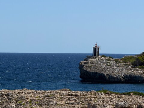 Lighthouse structure standing on a rugged mediterranean cliff, guiding ships navigating the deep blue sea under a clear summer sky in manacor, balearic islands, spain