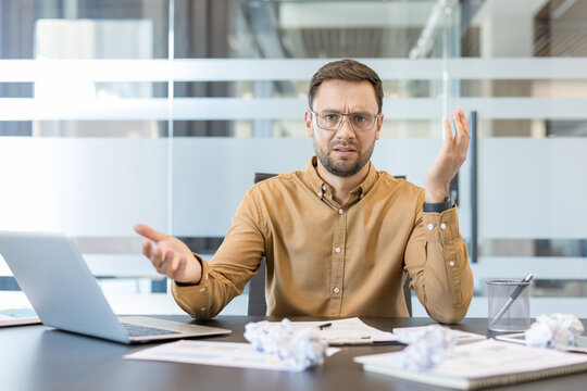 Work stress and burnout, frustrated businessman gesturing at desk with laptop and crumpled paper in office. Professional employee struggling with deadline pressure and heavy workload.