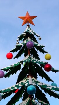 Colorful stylized christmas tree street decoration with a star on top and baubles on its snowy branches, rotating against a clear blue sky, celebrating the festive winter holiday season