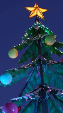 Close up of a contemporary urban christmas tree adorned with a glowing star and colorful baubles. Illuminated by a dynamic sequence of festive. Multicolored led lights against a deep blue evening sky